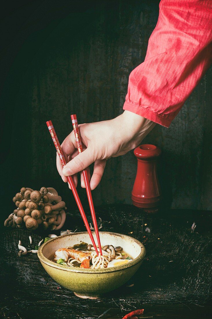 Female hand in red shirt take by chopsticks noodles from asian ramen soup with shrimp, onion, sliced egg and mushrooms