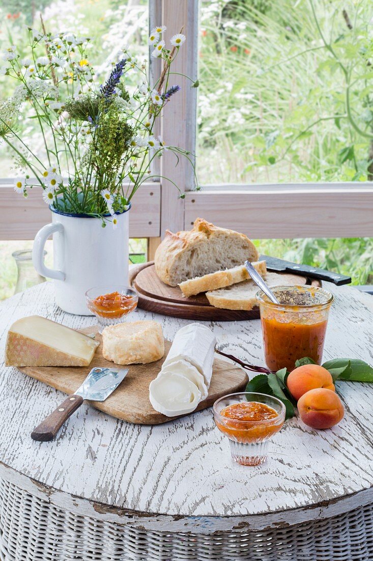 Cheese platter, rustic bread and chutney on white wooden table with flower bouquet and wooden boards