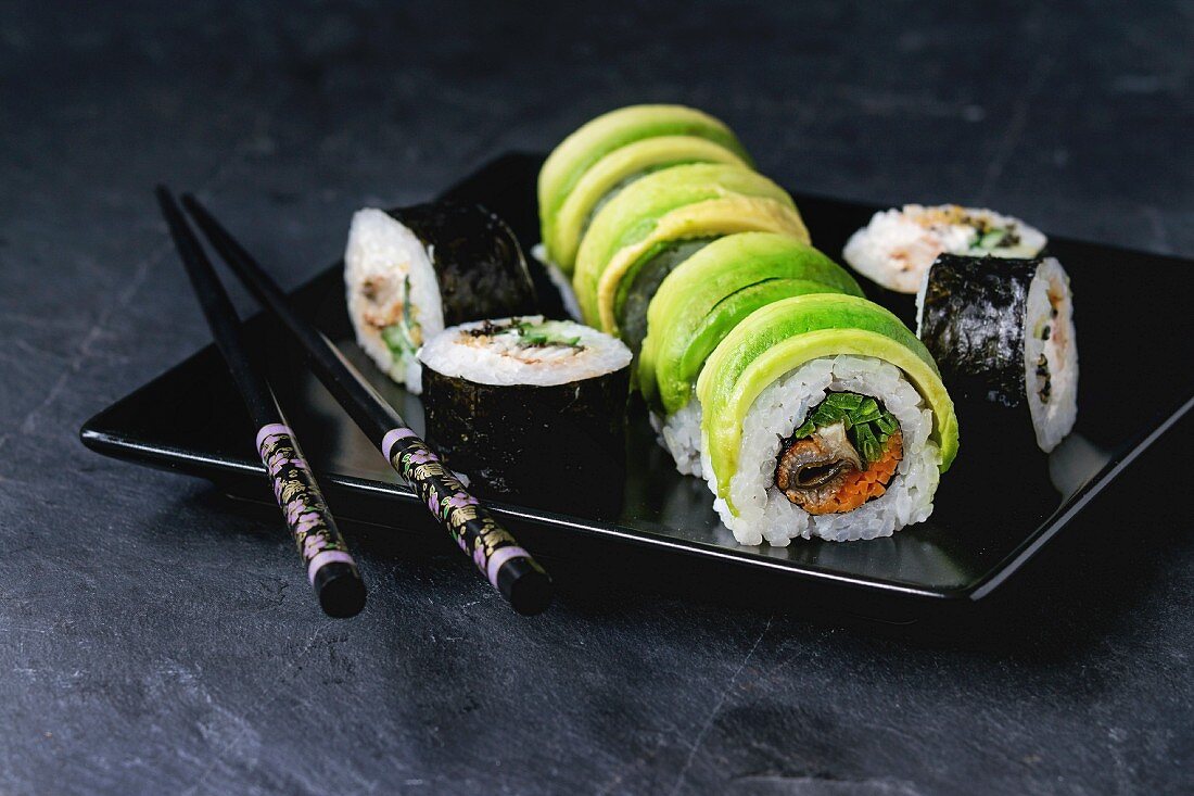Sushi set with avocado rolls served with chopsticks on square plate over black stone slate