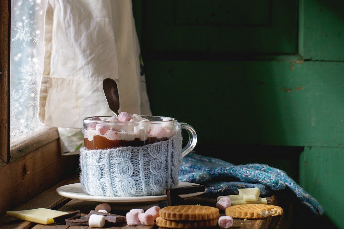 Heiße Schokolade mit Marshmallows in Glastasse mit gestrickter Husse, daneben Cookies und Fäustlinge