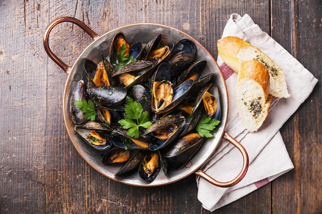 Mussels in copper cooking dish and French Baguette with herbs on wooden background