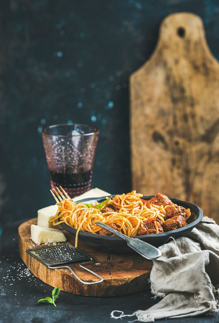 Spaghetti with meatballas, basil and parmesan cheese in black plate and red wine in glass over rustic wooden board
