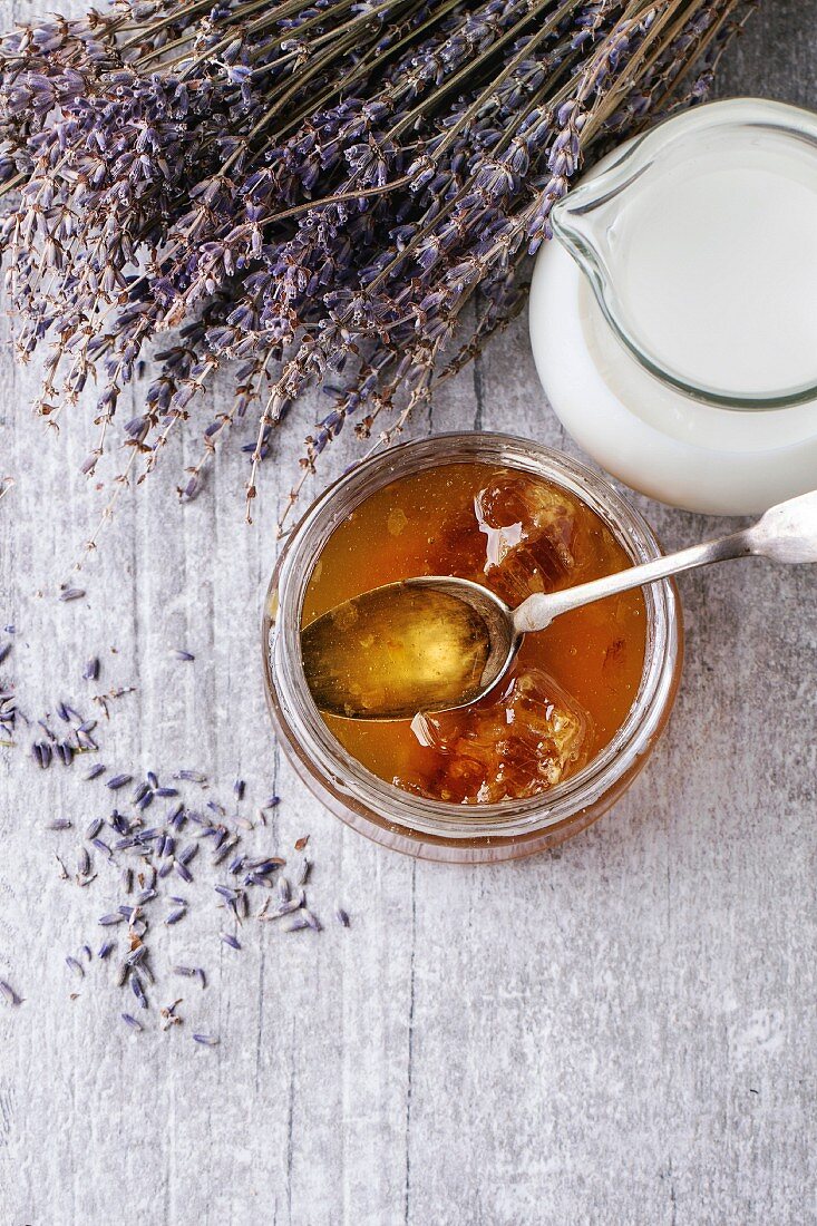 Open glass jar of liquid honey with honeycomb and silver spoon inside, glass jug of milk and bunch of dry lavender