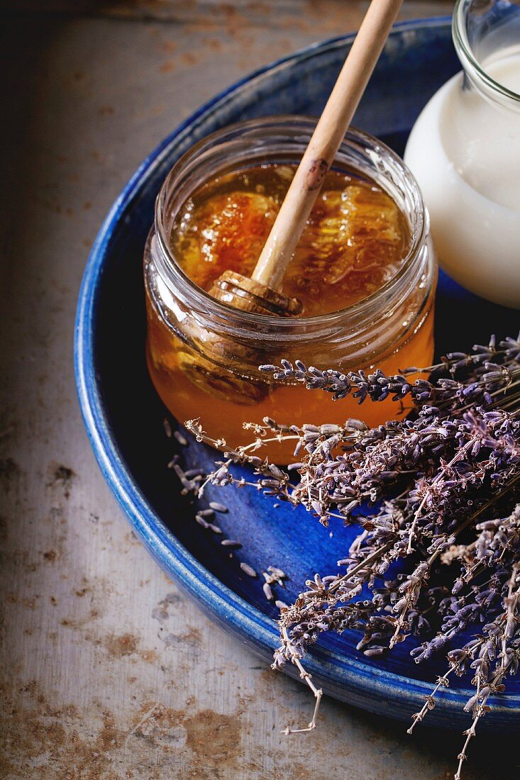 Open glass jar of liquid honey with honeycomb and honey dipper inside, milk and dry lavender