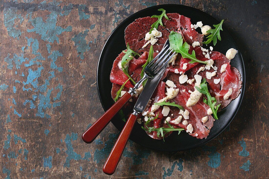 Beef carpaccio on black plate with mustard and parmesan sauce, cheese and arugula
