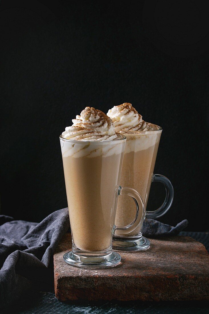 Pumpkin spicy latte with whipped cream and cinnamon in two glasses standing on clay board