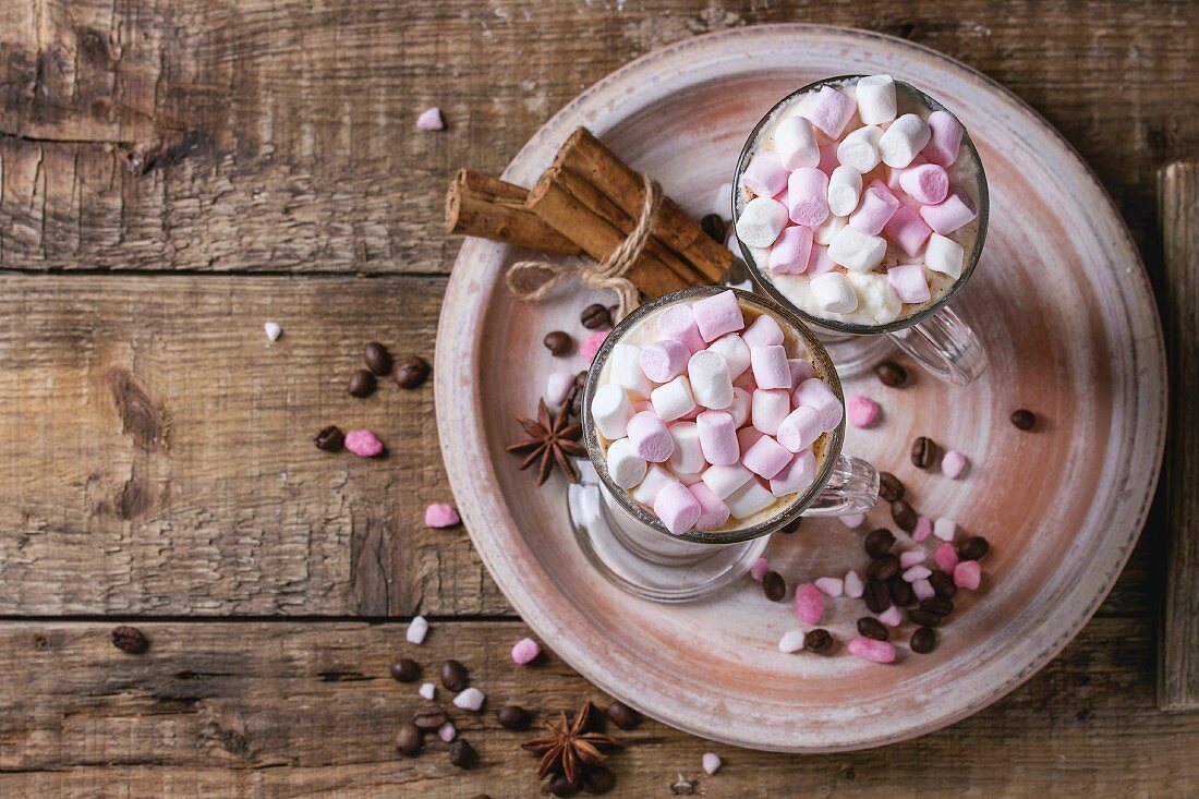 Two glasses of cafe latte with pink marshmallow standing on ceramic plate with spices, coffee beans and pink sugar
