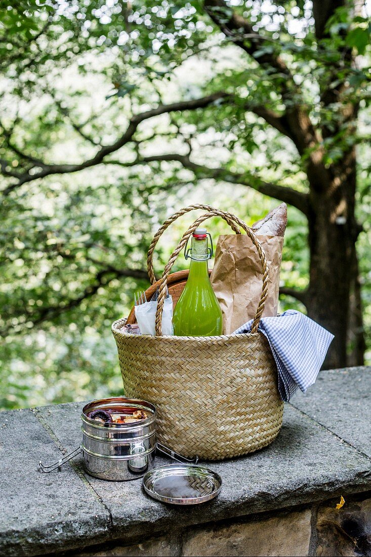 Picnic basket on wall with lemonade bottle, lunch box, artisan bread in a paper bag, and picnic blanket