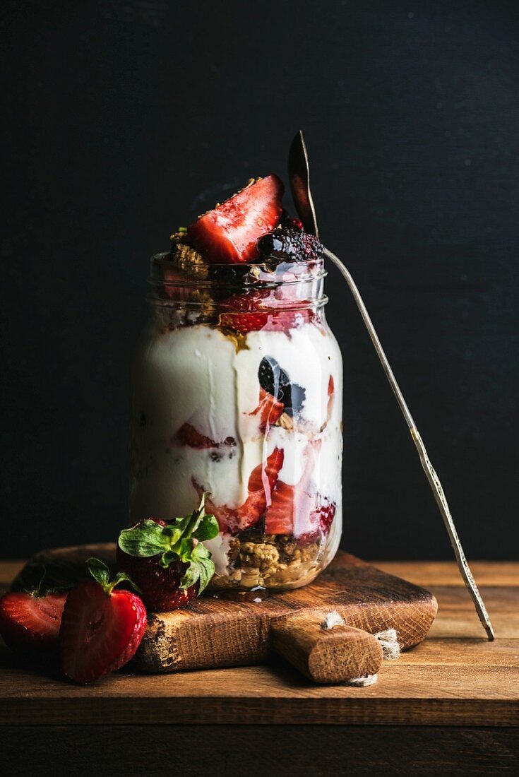 Granola-Müsli mit Joghurt, Erdbeeren und Maulbeeren in Glas vor schwarzem Hintergrund