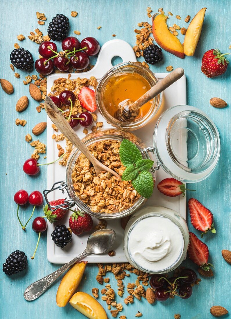 Oat granola in open glass jar, yogurt, fruit, berries, honey and mint on white ceramic board
