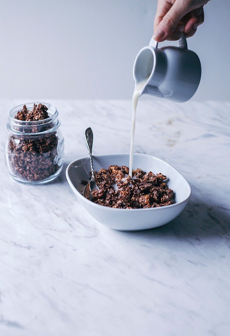 Woman pouring almond milk into a bowl of homemade chocolate granola with oats, nuts and seeds
