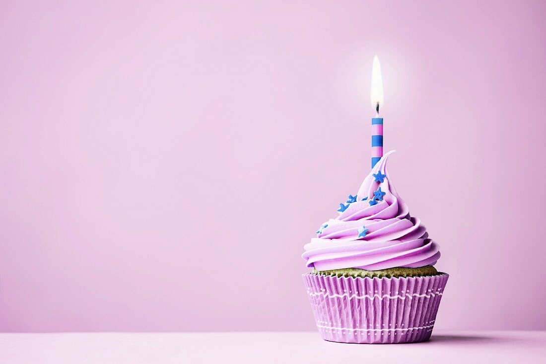 Lilafarbener Geburtstags-Cupcake mit brennender Kerze vor violettem Hintergrund