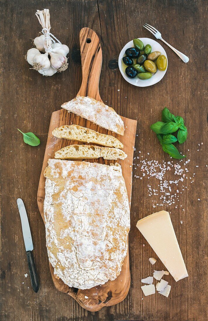 Ciabatta-Brot auf Holzbrett daneben Knoblauch, Oliven, Basilikum und Parmesan