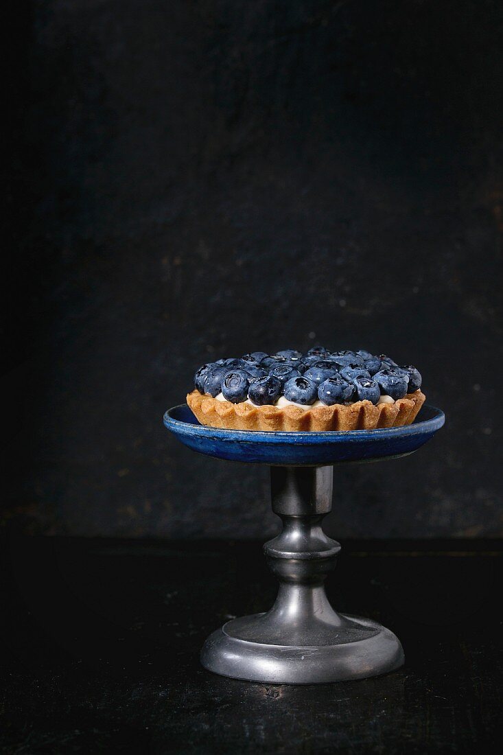 Lemon tartlet with fresh blueberries, served on vintage cake stand over black background