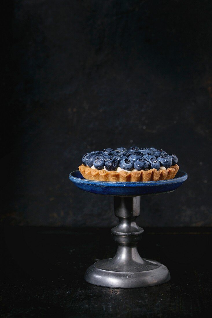 Zitronentörtchen mit Blaubeeren auf Vintage-Kuchenständer vor schwarzem Hintergrund