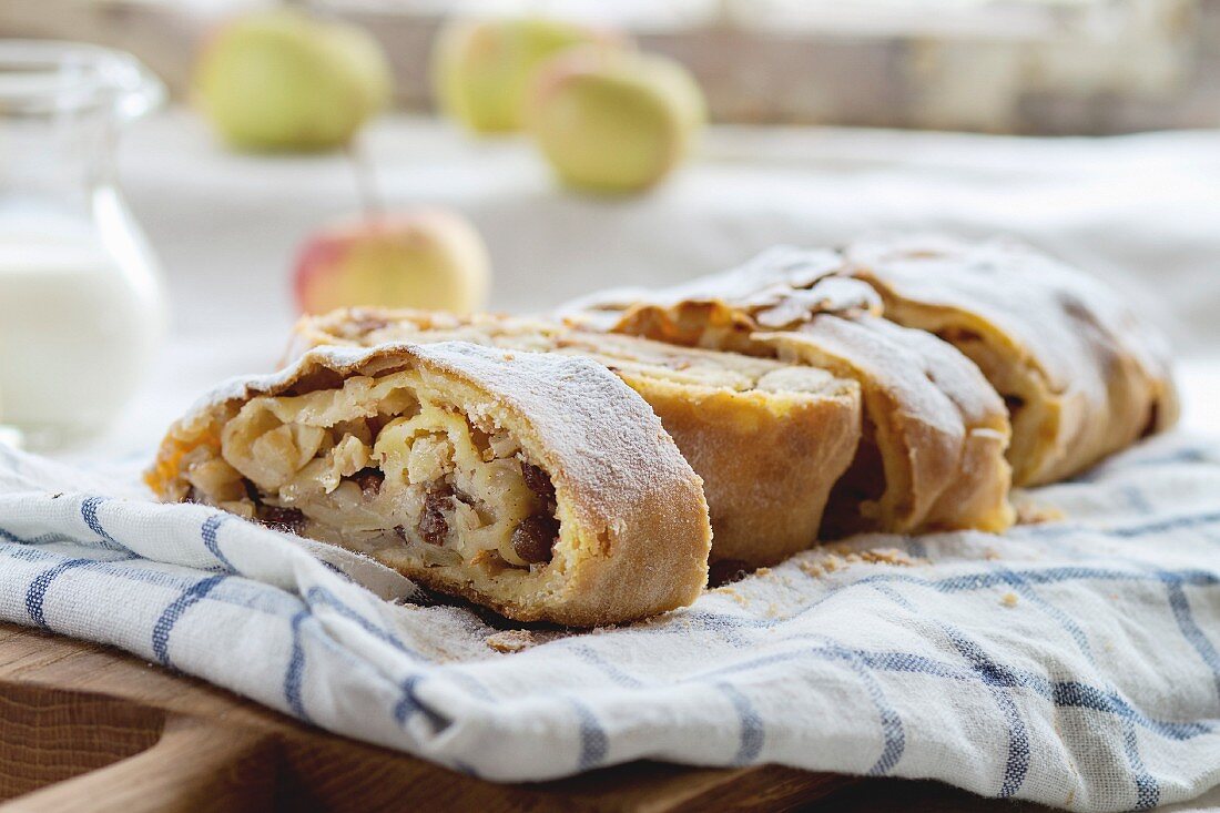 Sliced fresh baked homemade apple strudel over towel on kitchen table with jug of milk and apples