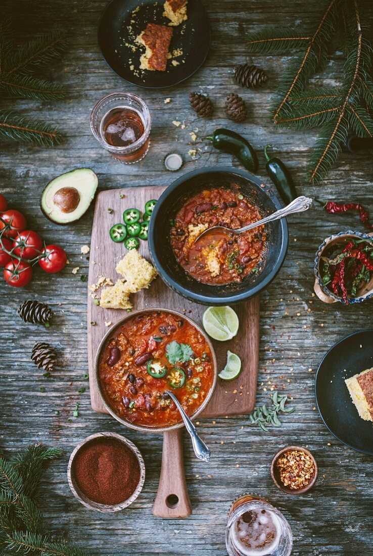 Two bowls of Turkey and Bean chili surrounded with two glasses of beer, corn bread, tomatoes, chili powder