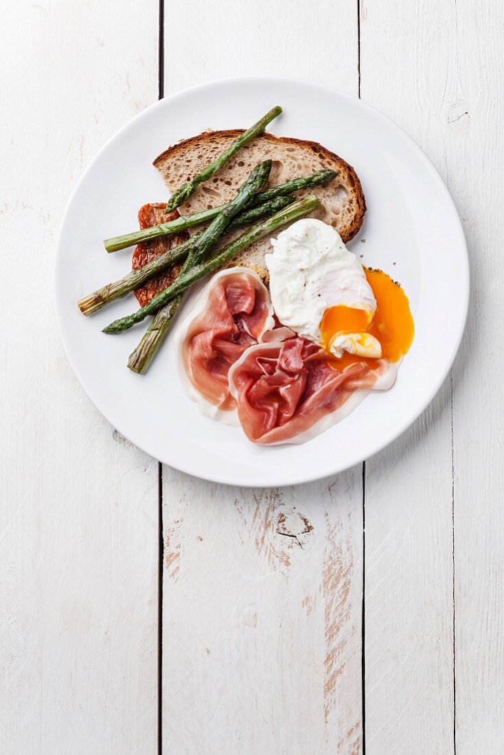 Breakfast with poached egg, parma and asparagus on white wooden background