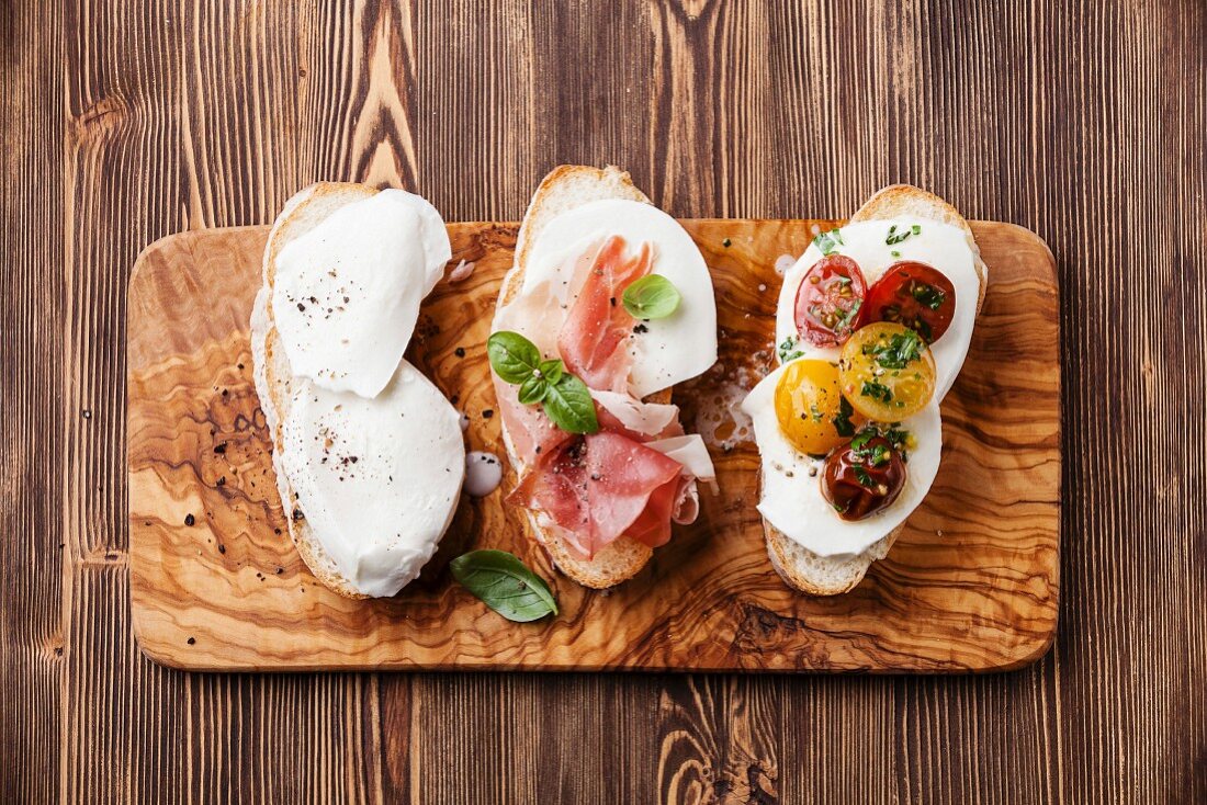 Three bruschettas with mozzarella cheese, ham and roasted tomatoes on ciabatta bread on wooden background