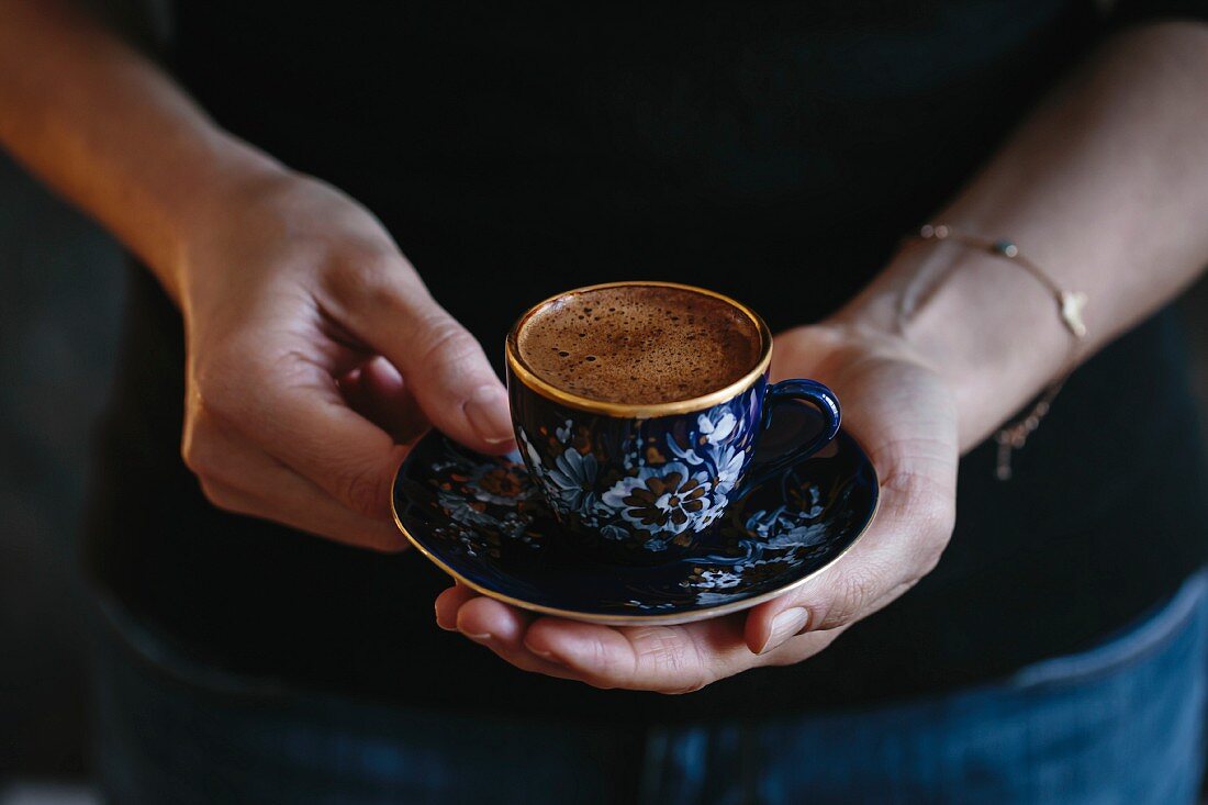 A woman is holding a cup of Turkish coffee with foam on top (with both hands)