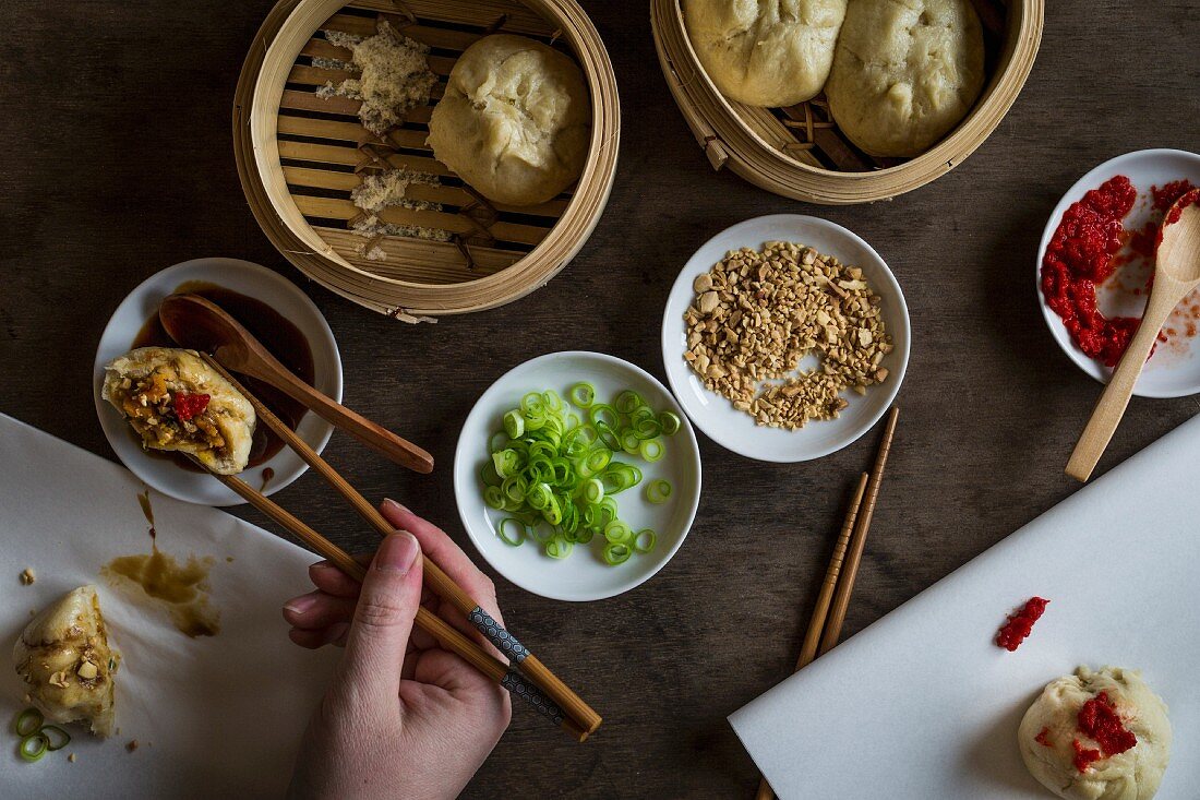 Woman s hand with chopsticks dipping stuffed dumpling in sauce on wooden tabletop with bamboo steamers