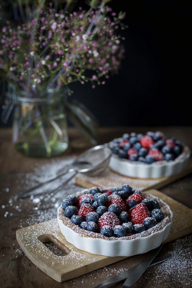 Berry tartetelets with icing sugar in baking dishes on wooden plates with bouquet of flowers in background