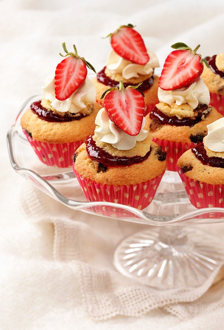 Several fresh strawberries and cream cupcakes on a glass cake stand with cream coloured naplin and cloth