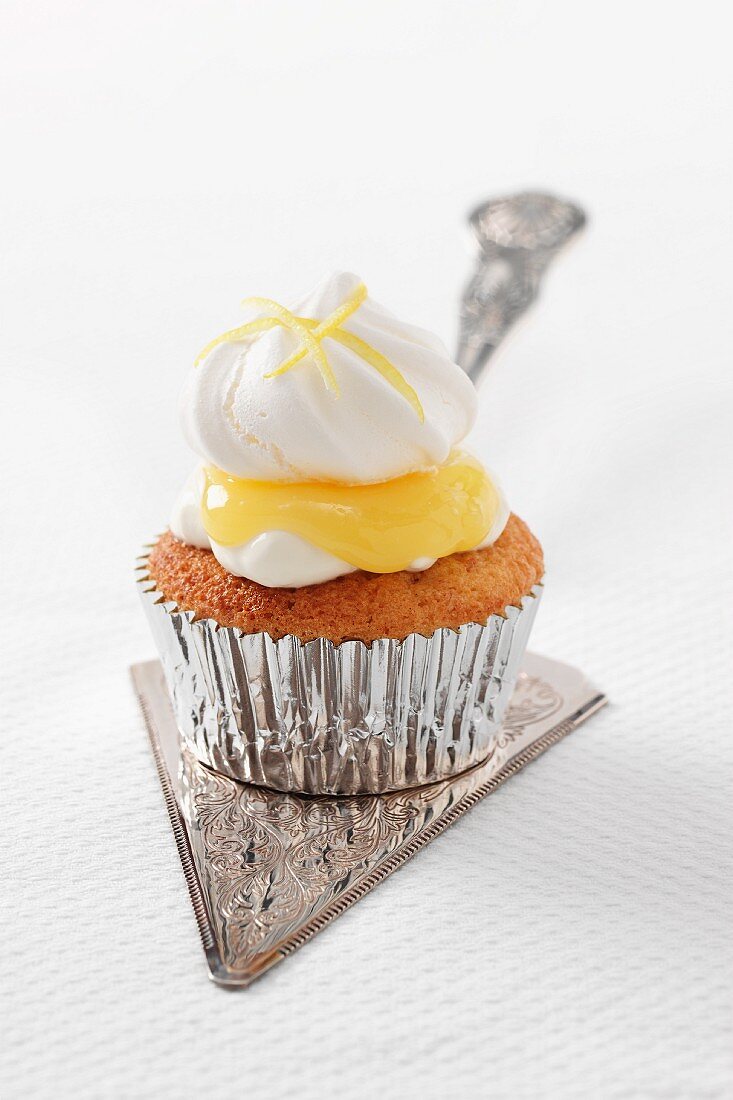 A single Lemon meringue cupcake with lemon curd in a silver case sitting on a silver cake slice