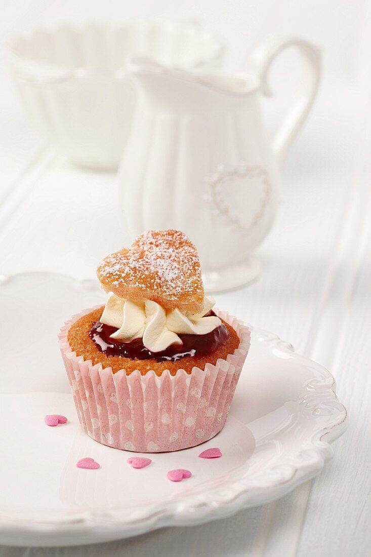 A sweet heart cupcake on a white plate with white milk jug and sugar basin in background
