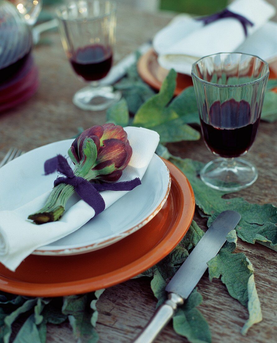 Purple globe artichoke on white serviette and orange plate on wooden table with large artichoke leaves and glasses of red wine