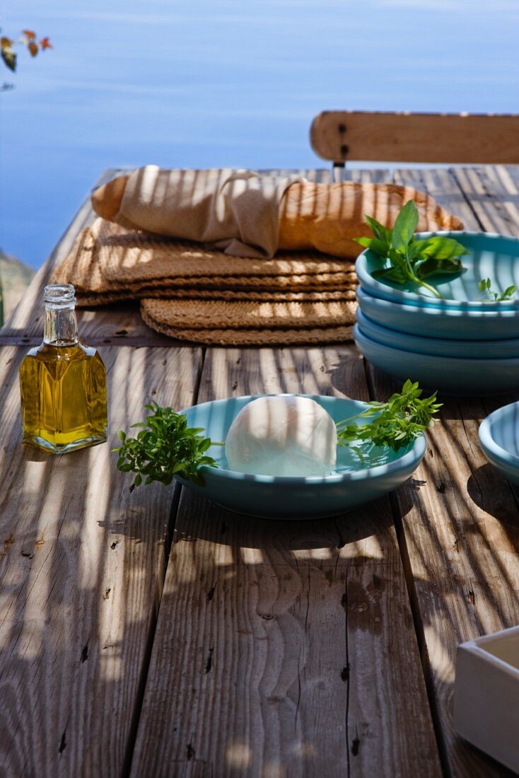 Rustikaler Holztisch am Meer mit blauen Tellern, Brot, Käse und Olivenöl