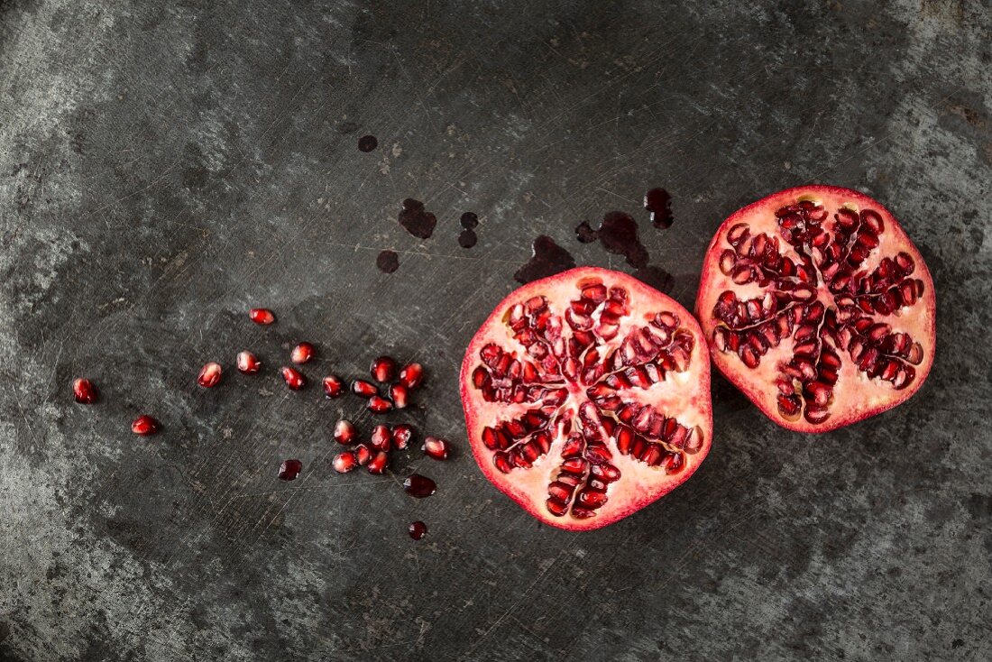 Two halves of a pomegranate and seeds and juice on a grey metal surface