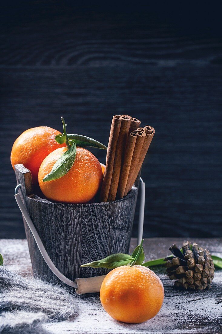 Wooden bucket with tangerines and cinnamon sticks over wooden background with snow and cone