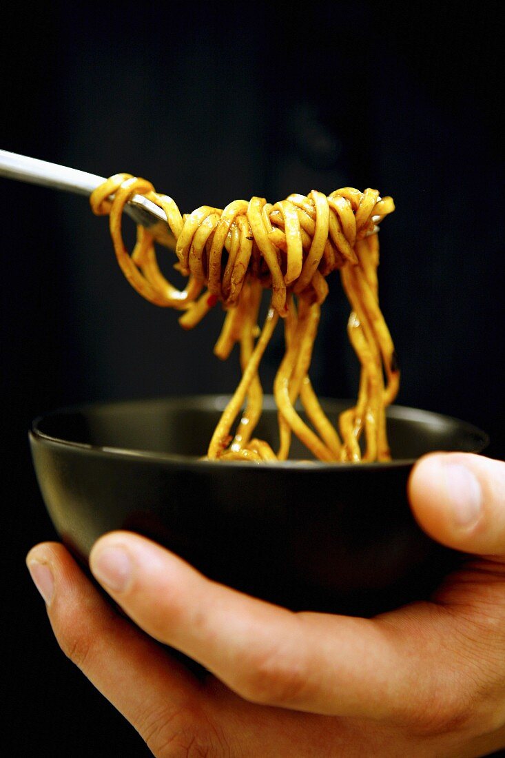 Person eating noodles out of a black ceramic bowl using a fork, black background.