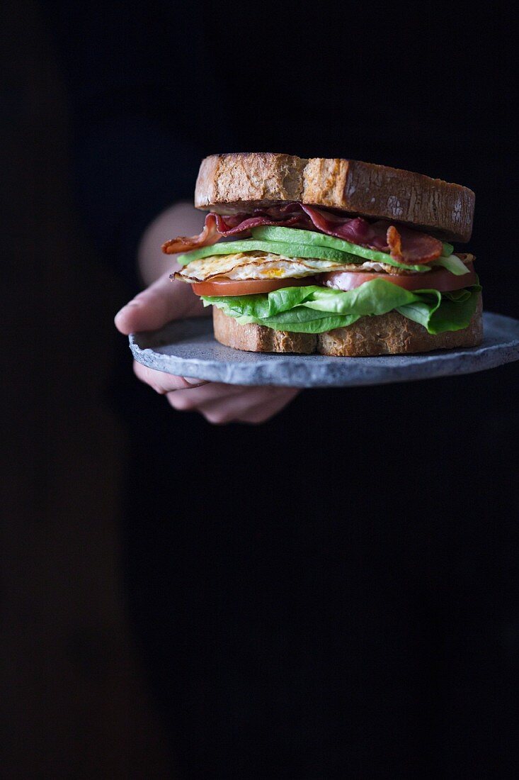 Hand hält Sandwich mit Spiegelei, Bacon, Avocado, Tomaten und Salat