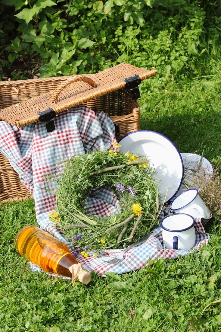 Picnic basket, blanket, enamel crockery and wreath of wildflowers on green lawn