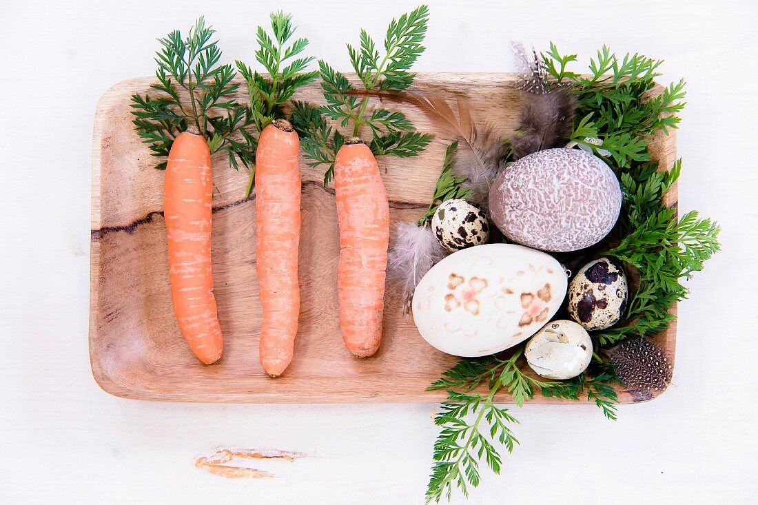 Carrots, carrot leaves, feathers and various decorated eggs in wooden dish