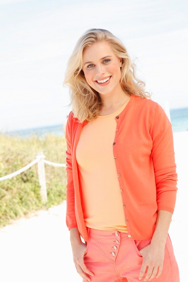 Blonde Frau in T-Shirt, Cardigan und Hose in verschiedenen Orangetönen am Strand