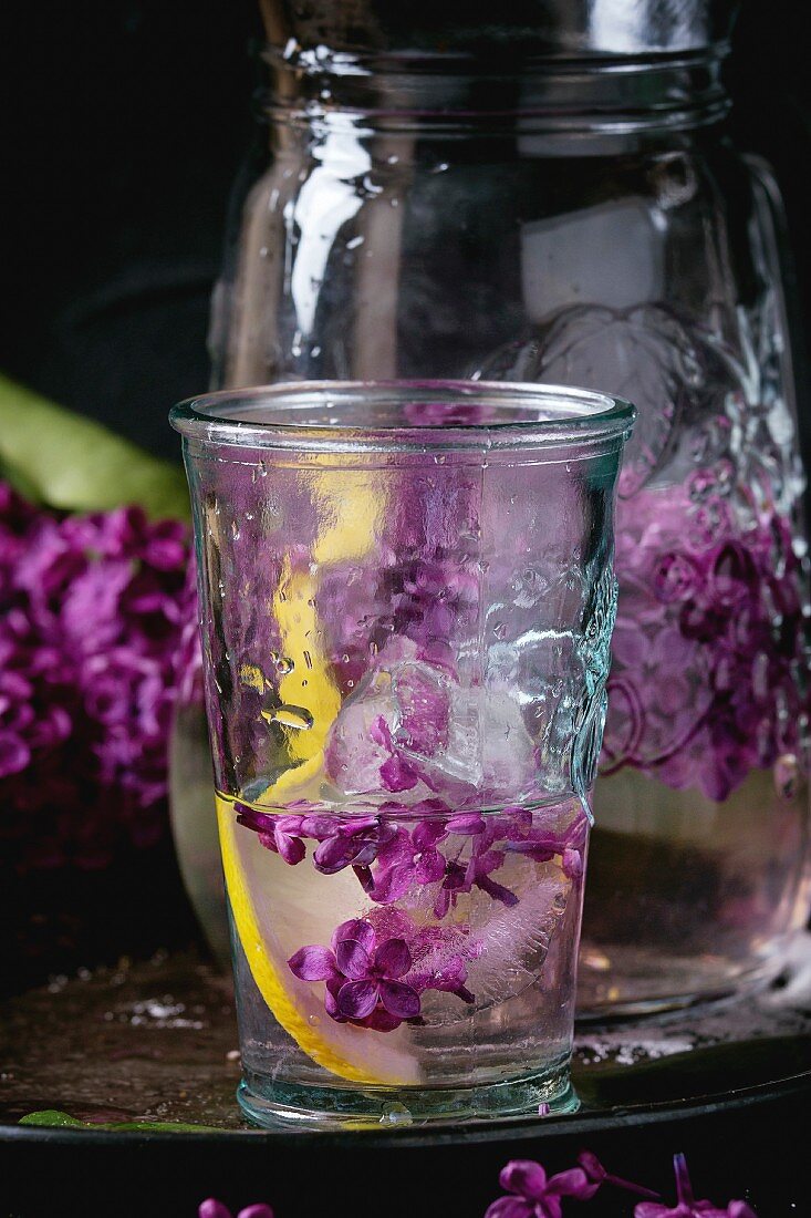 Glass and pitcher of lilac lemonade water with lemon, ice cubes with lilac flowers and lilac branch