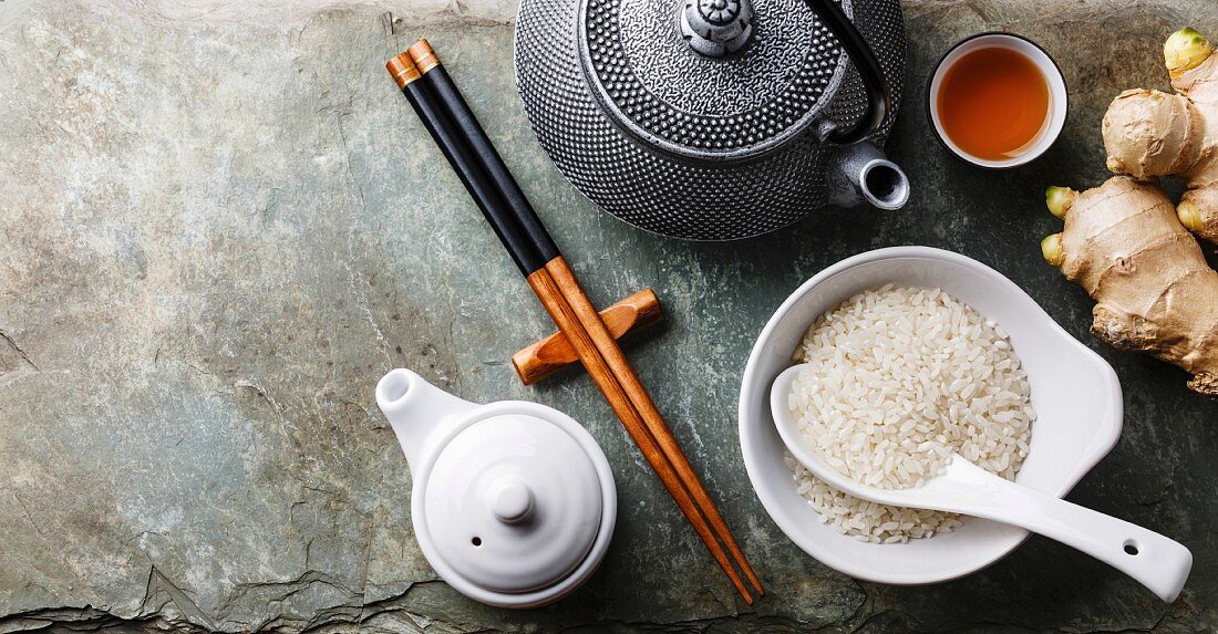 Raw white rice, green tea teapot, soy sauce and sushi chopsticks on gray stone slate background