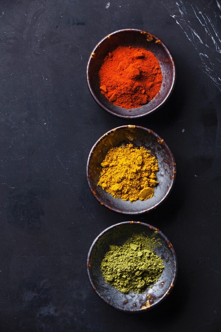 Paprikapulver, Curry und Matcha-Teepulver in Schälchen vor dunklem Hintergrund