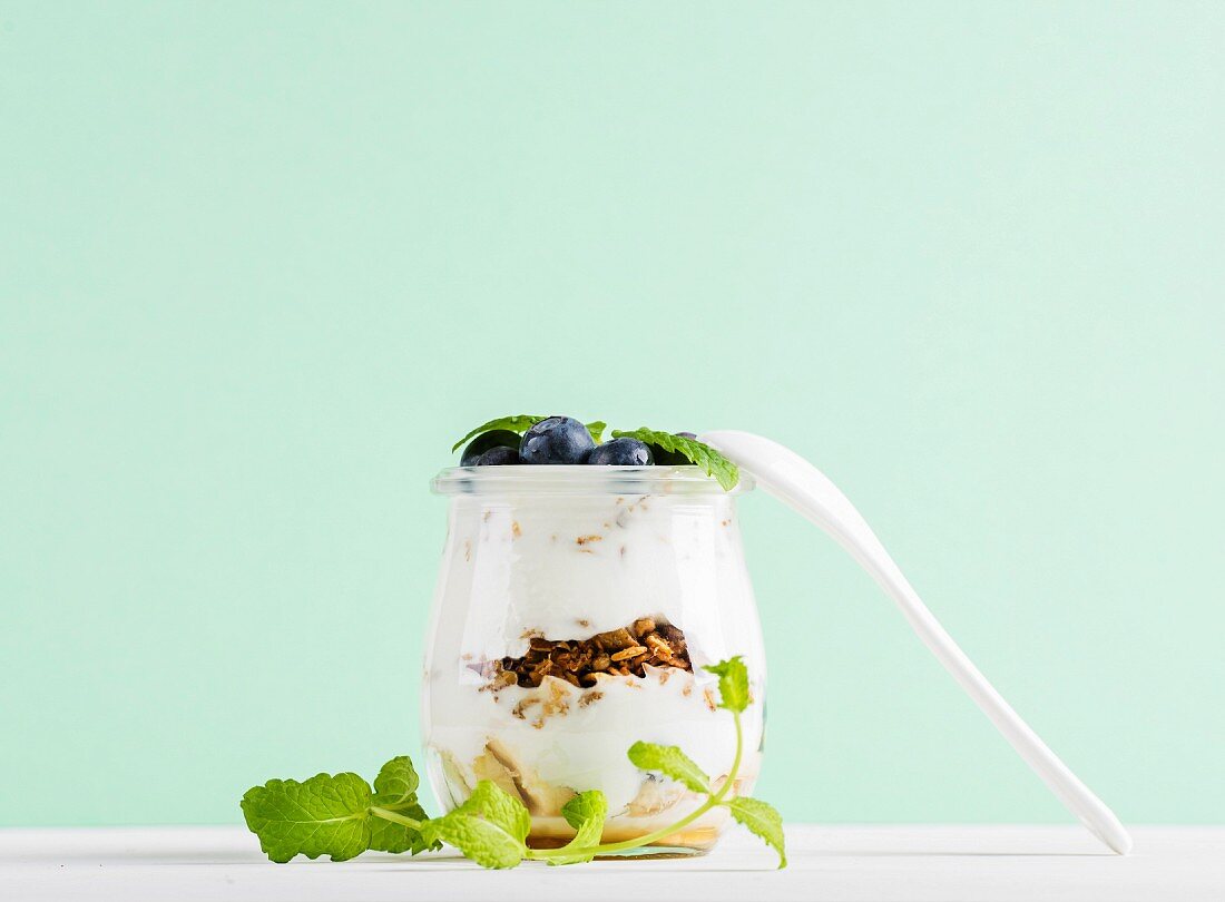 Yogurt oat granola with fresh berries and mint leaves in glass jar on grey concrete textured backdrop