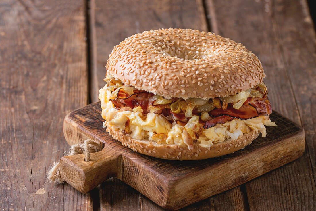 Bagel with bacon, scrambled eggs and fried onion on small wooden chopping board