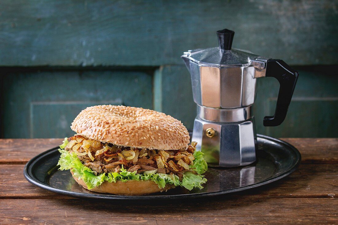 Bagel with stew beef, fresh salad and fried onion served on vintage metal tray with coffee pot