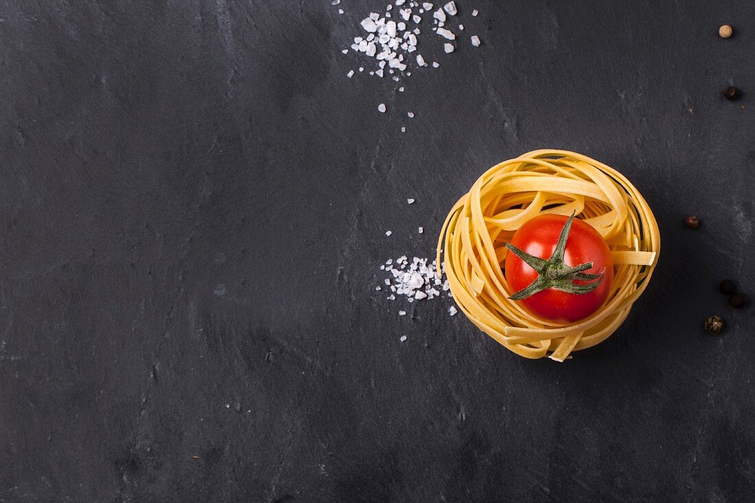 Top view on dry pasta and fresh cherry tomato served with sea salt on dark grey slate background