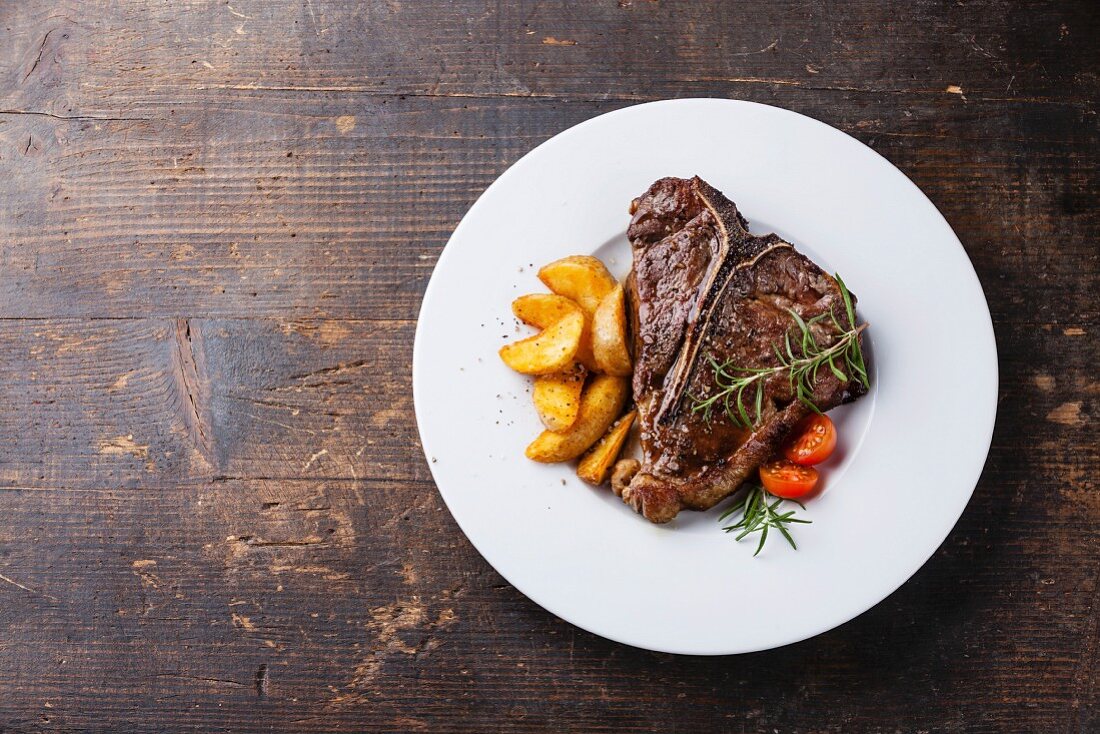 T-Bone Steak with roasted potato wedges on white plate on wooden background