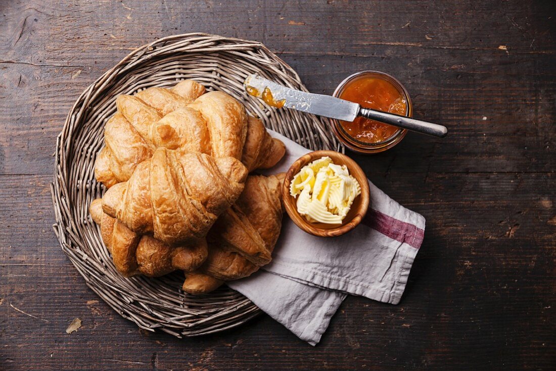Croissants with butter and jam in wicker tray on dark wooden background