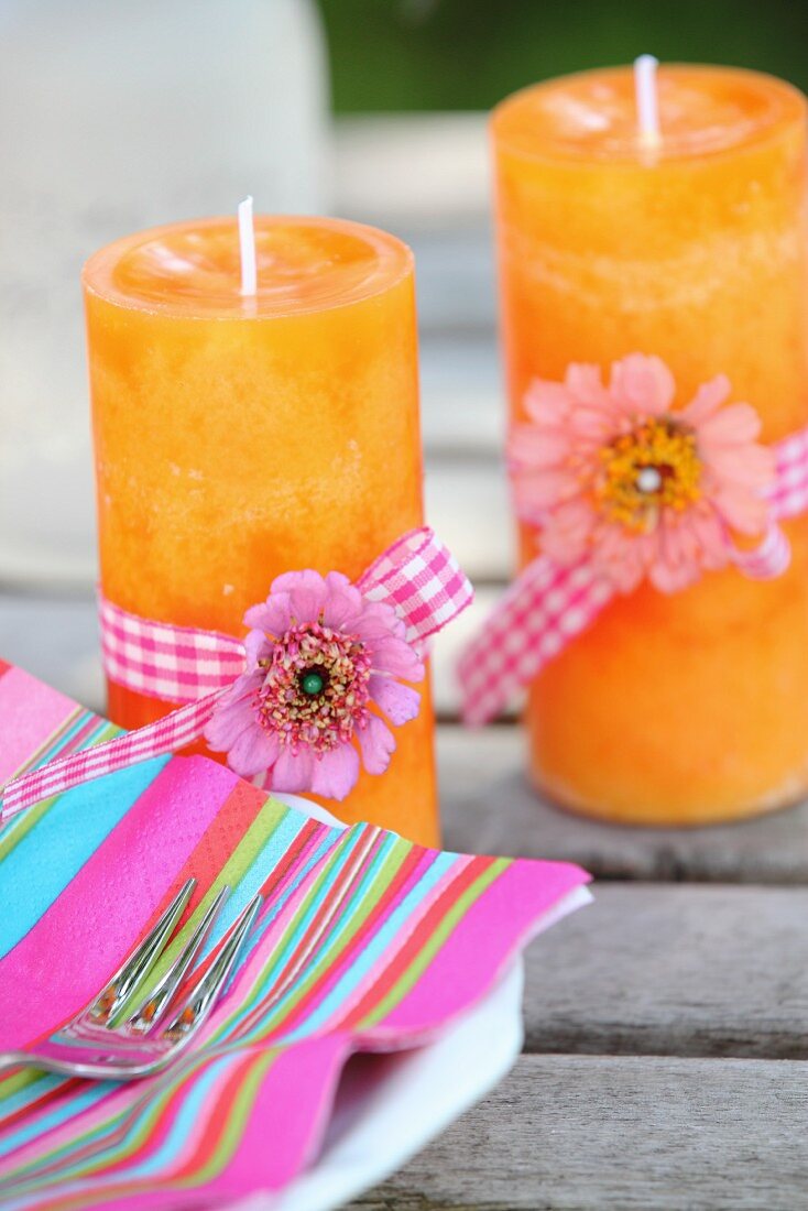 Pinkfarbener Streifenserviette und zwei orangefarbenen Kerzen mit Blüten und Geschenkbändern dekoriert