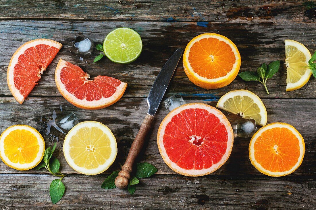 Scheiben von Zitrone, Limette, Orange und Grapefruit, mit Minze, Eis und Vintage-Messer auf Holzuntergrund