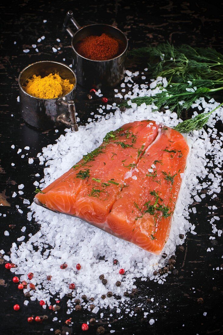 Piece of salted salmon, served with dill and spices on sea salt over black wooden table